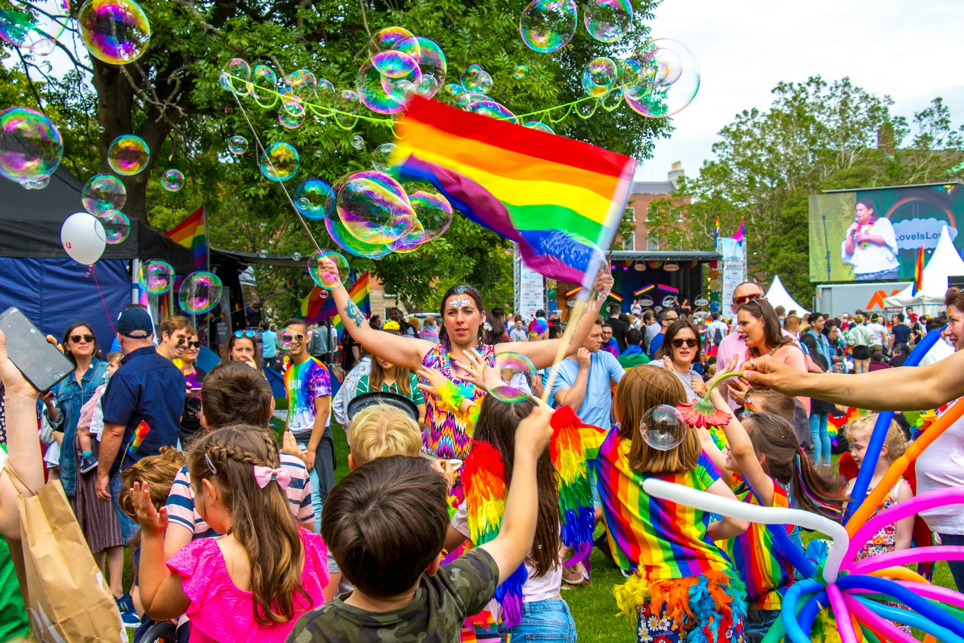 A mixed group of revelers including adults and children dressed in rainbow colors celebrate Pride Festival in Dublin in June, flying flags and blowing bubbles